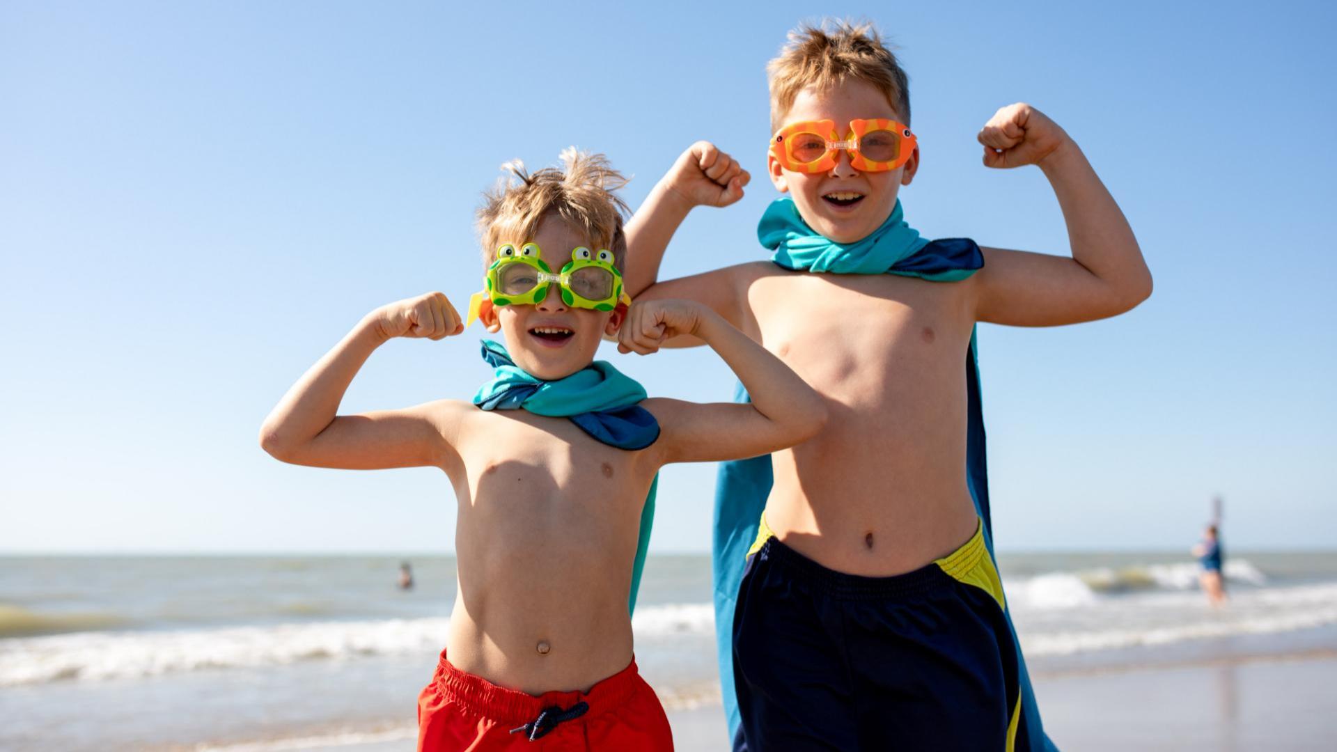 Due bambini in costume da bagno e occhialini giocano a fare i supereroi sulla spiaggia.